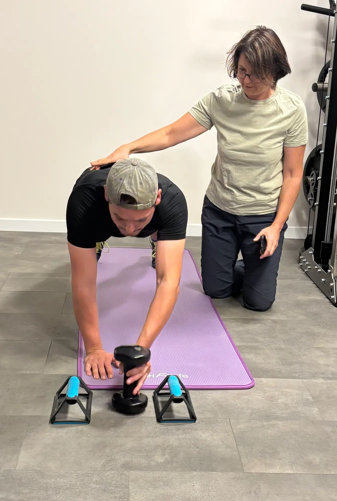 Coach sportive corrigeant la posture d'un client en gainage sur tapis, un haltère à la main entre deux poignées de pompes